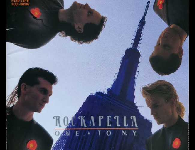 Cover of 'Rockapella One: To NY' which features images of Rockapella popping out of the four corners of the cover with an image of the empire state building in the background. Starting at the top left corner and going clockwise, we see Elliot, Barry, Scott, and Sean all wearing matching dark coats over matching dark tshirts. They are mostly in the dark with a bright red flower pinned to each of their chests seemingly more illuminated.